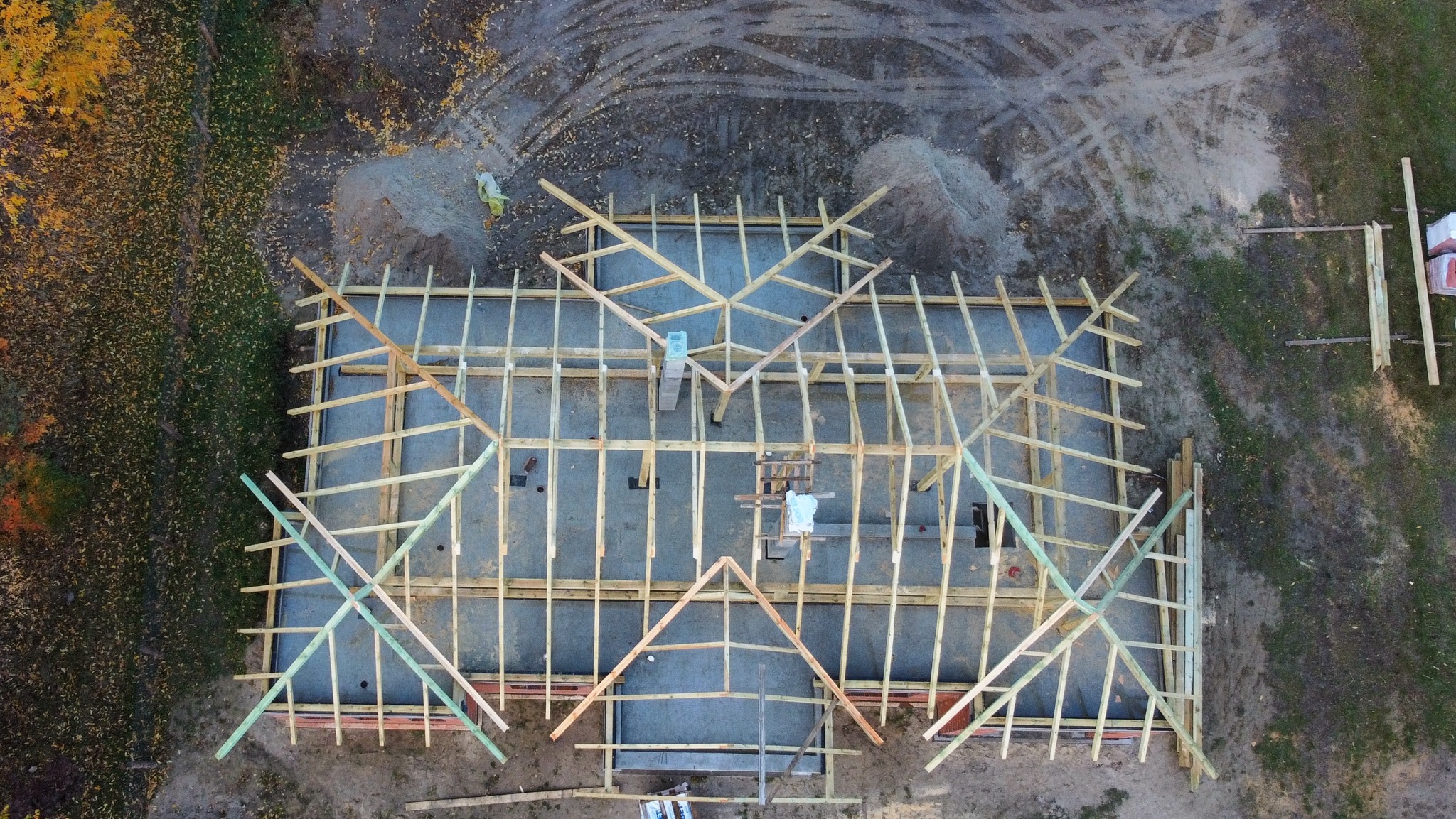 Widok z drona na budowę domu w trakcie montażu drewnianej konstrukcji dachu, widoczne elementy więźby dachowej i podkład, w tle jesienne drzewa i plac budowy.