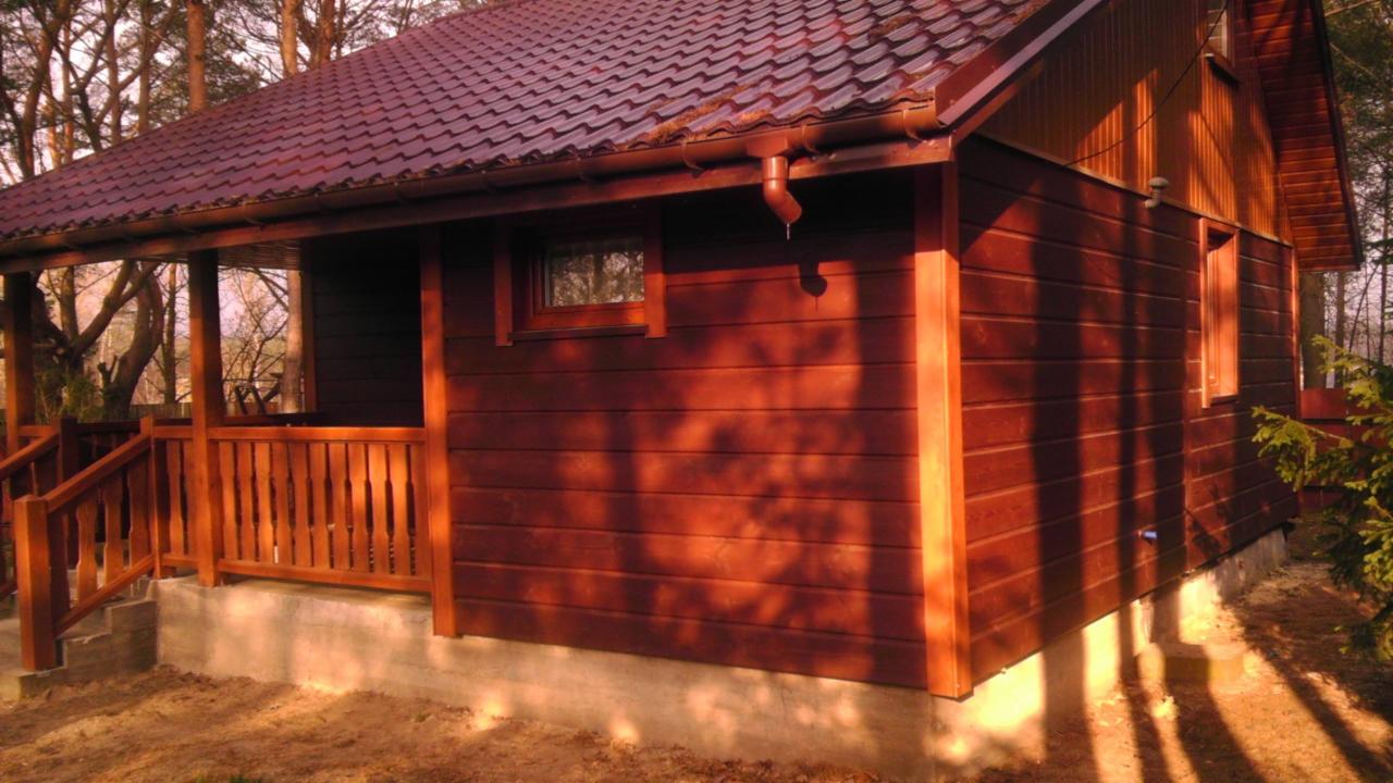 Zewnętrzny widok brązowego, drewnianego domku letniskowego z gontowym dachem, werandą i betonową podmurówką, otoczonego drzewami.