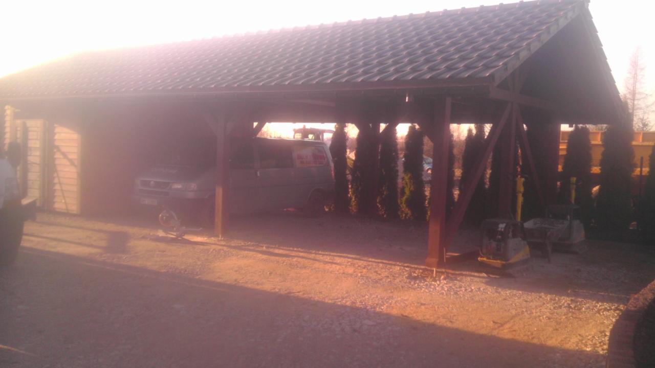 Zadaszenie z drewnianą konstrukcją i dachówką ceramiczną, pod którym stoi bus i sprzęt budowlany, w tle rząd tuj.