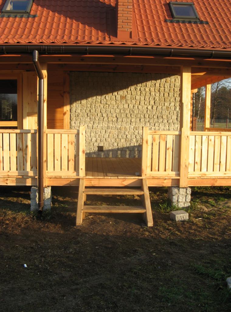 Surowa drewniana konstrukcja domu z elementami kamiennej elewacji, widoczne schody wejściowe i fragment dachu z dachówką ceramiczną.