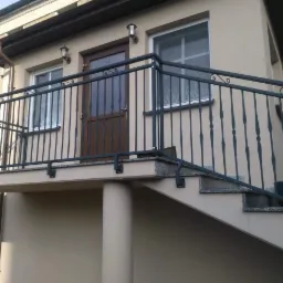 Zewnętrzna, metalowa balustrada z dekoracyjnymi elementami na balkonie i schodach wejściowych do domu z beżową elewacją.