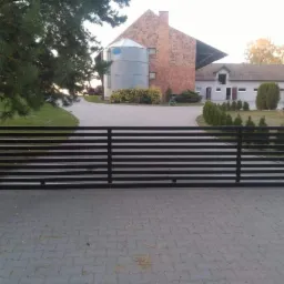 Nowoczesna, pozioma brama przesuwna w kolorze czarnym, z widocznymi ceglanymi słupkami i brukowaną nawierzchnią podjazdu, w tle budynki gospodarcze i silos.