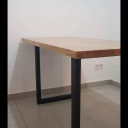 Stół z blatem z jasnego drewna i czarnymi, metalowymi nogami w kształcie prostokątów, ustawiony w rogu pomieszczenia z białymi ścianami i jasną podłogą z płytek.