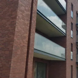Nowoczesny budynek wielorodzinny z elewacją z czerwonej cegły, przeszklonymi balkonami i metalowymi balustradami, widziany pod kątem z dołu.
