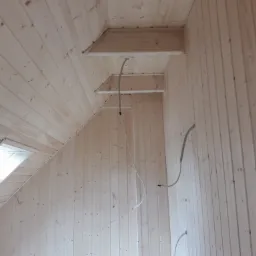 Wnętrze poddasza wykończonego boazerią z widocznym oknem dachowym i przewodami instalacji elektrycznej.