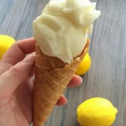 Ręka trzyma wafel z cytrynowymi lodami na tle trzech cytryn na szarym drewnianym stole.