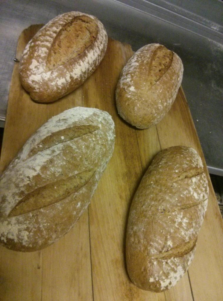 Cztery bochenki chleba rustykalnego, posypane mąką, ułożone na drewnianej desce do krojenia, widoczne z góry pod kątem.