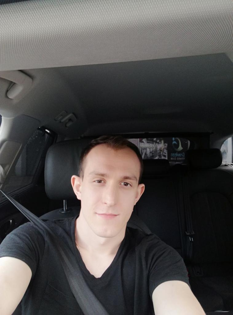 Młody mężczyzna w czarnej koszulce, zapięty pasami w samochodzie, spogląda w obiektyw. Widoczne wnętrze pojazdu i szyba.