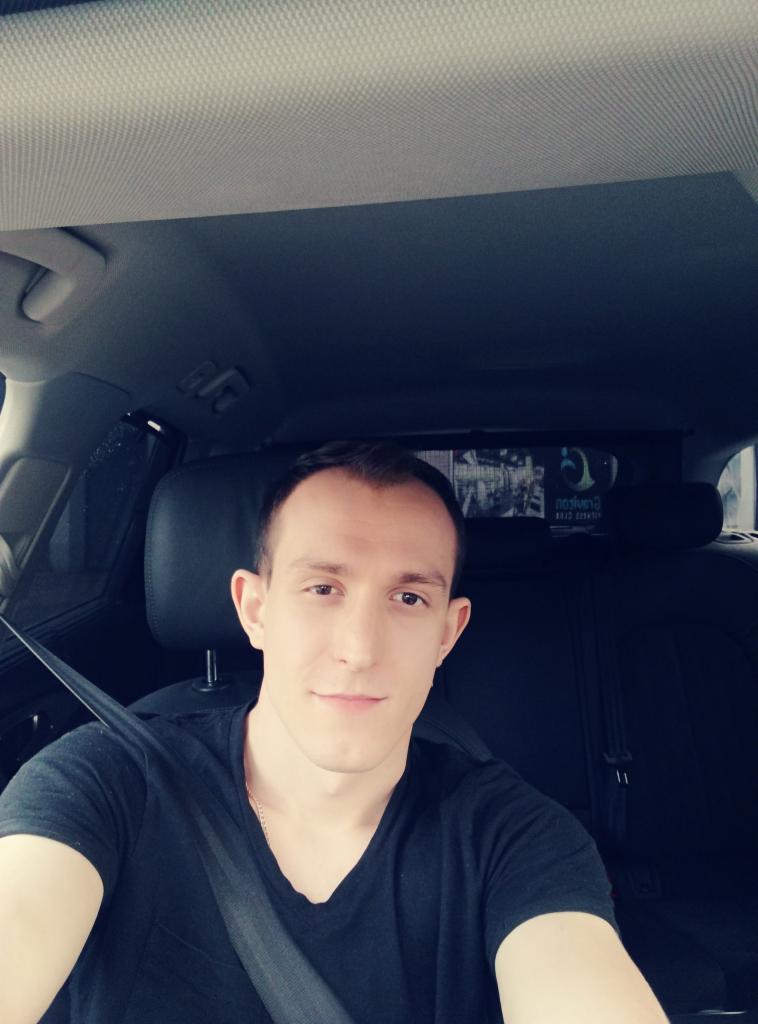 Mężczyzna w czarnej koszulce zapięty pasem w samochodzie z czarną tapicerką, widoczny szyberdach i zagłówek tylnego siedzenia.