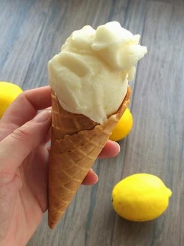 Ręka trzyma wafel z cytrynowymi lodami na tle trzech cytryn na szarym drewnianym stole.