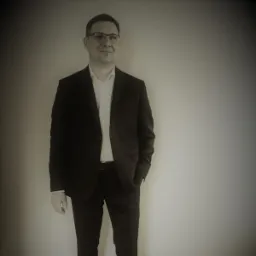 Portret mężczyzny w garniturze i okularach, z ręką w kieszeni, stojącego na tle szarej ściany. Uśmiecha się delikatnie, patrząc w bok.