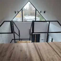 Wnętrze domu z antresolą i metalową balustradą o geometrycznym wzorze, widok z góry na drewnianą podłogę antresoli, trójkątne okno w szczycie dachu i minimalistyczne oświetlenie.