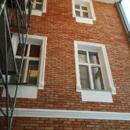 Ściana wewnętrzna wyłożona czerwoną cegłą klinkierową z białymi obramowaniami okiennymi, widoczne rusztowanie z lewej strony.