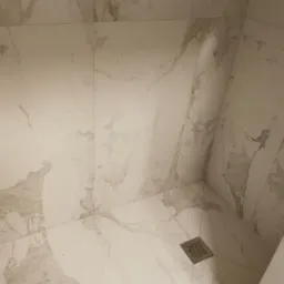 Wykończona kabina prysznicowa z białych płytek imitujących marmur, z odpływem liniowym w rogu.