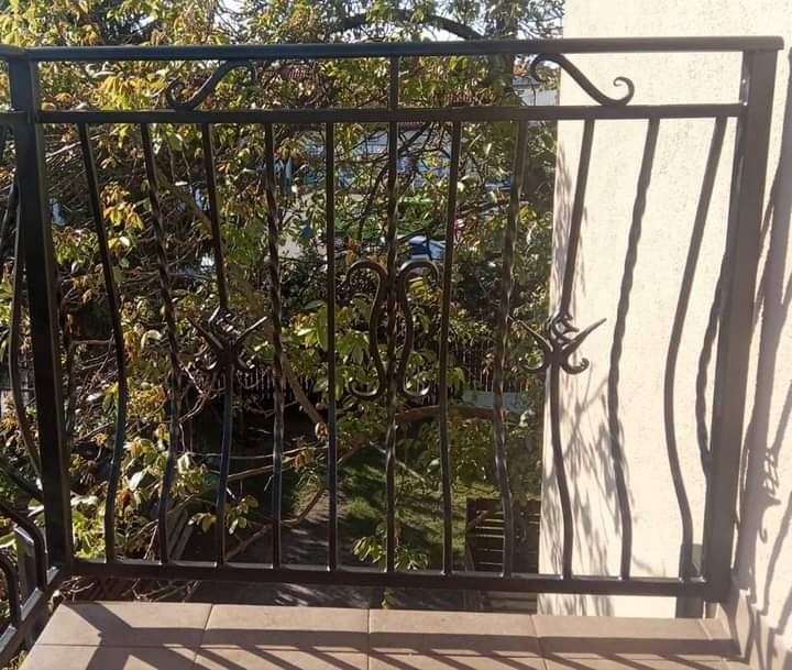 Czarna, kuta balustrada balkonowa z dekoracyjnymi elementami roślinnymi, widok z góry na zielone drzewa w tle.