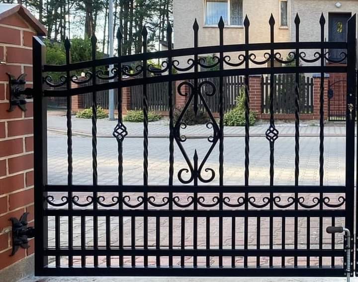 Czarna, metalowa brama wjazdowa z dekoracyjnymi elementami w kształcie spirali i serc, zamontowana na ceglanym słupku.