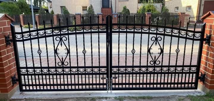 Czarna, metalowa brama wjazdowa z dekoracyjnymi elementami, zamontowana między ceglanymi słupkami, na tle domu i ogrodzenia.