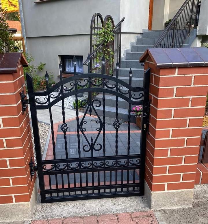 Czarna, kuta furtka z ozdobnymi elementami, osadzona między dwoma ceglanymi słupkami z daszkami, na tle domu z schodami.