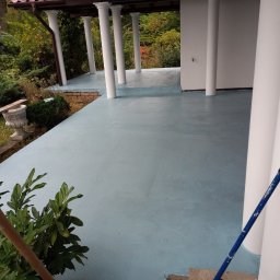 Usługi budowlane Marek Sulecki - Błękitna, świeżo pomalowana podłoga na zewnątrz budynku z kolumnami. Widoczne narzędzia malarskie i zieleń. Ujęcie z bliska, podkreślające gładką powierzchnię.