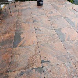 Usługi budowlane Marek Sulecki - Wykończony taras z płytek imitujących kamień w odcieniach brązu i szarości. Widoczny kubeł z zaprawą i poziomica. Częściowo widoczne krzesło i fragment elewacji.