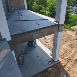 Usługi budowlane Marek Sulecki - Wykończony taras z szarych płytek, widok z góry. Na płytkach leżą narzędzia, obok stoi rower i przyczepka. W tle zieleń i budynek.