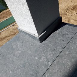 Usługi budowlane Marek Sulecki - Szara, kamienna posadzka tarasu z fugami, przylegająca do otynkowanej kolumny. W tle widoczny fragment terenu z zielonymi elementami konstrukcyjnymi.