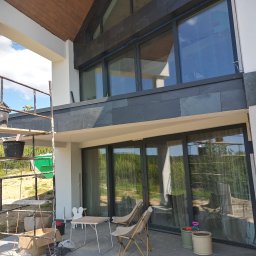 Usługi budowlane Marek Sulecki - Nowoczesny dom z szarą elewacją i dużymi oknami, widok z rusztowania. Elementy wykończeniowe z kamienia. Na tarasie stolik i krzesła.