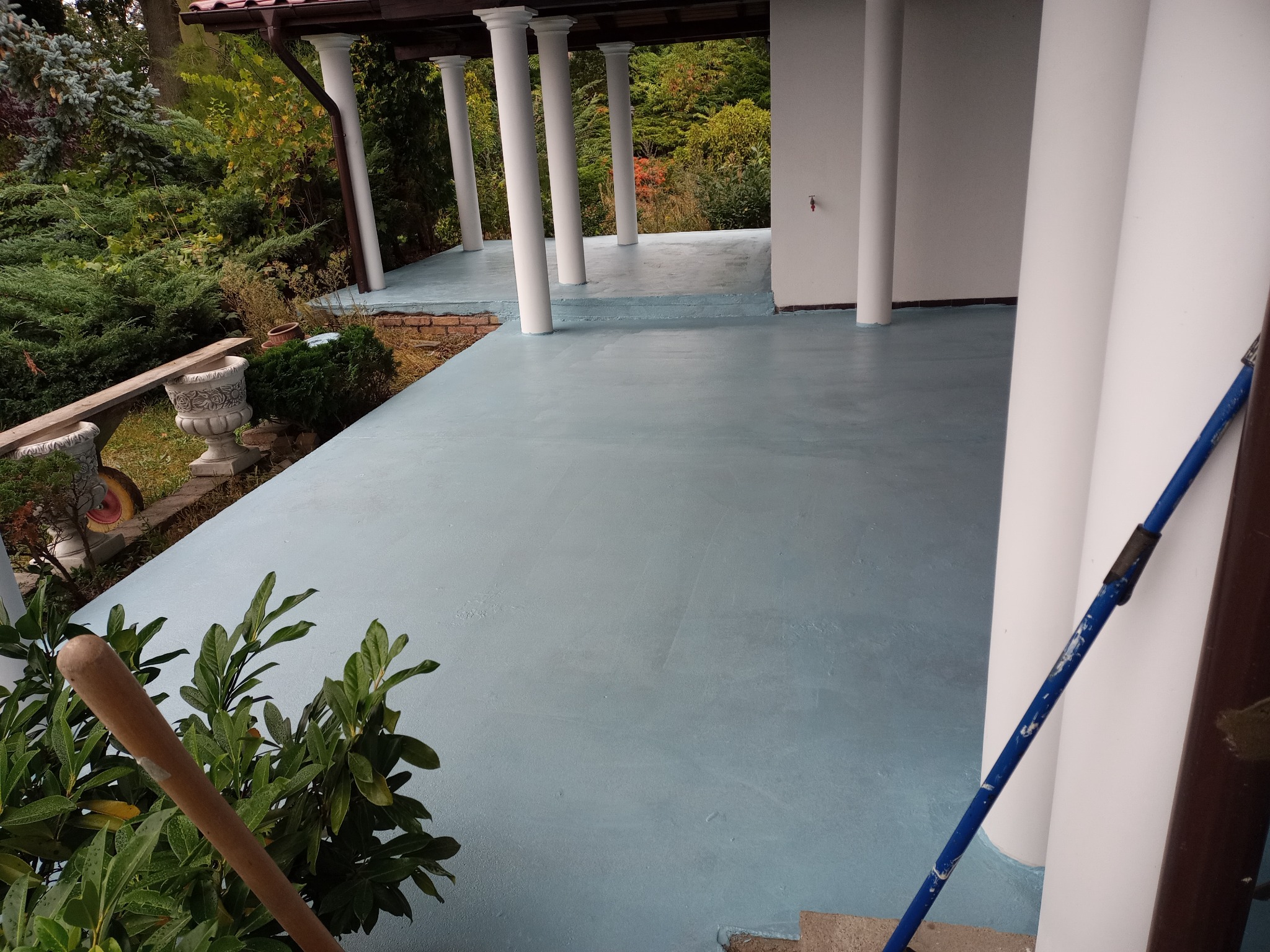 Błękitna, świeżo pomalowana podłoga na zewnątrz budynku z kolumnami. Widoczne narzędzia malarskie i zieleń. Ujęcie z bliska, podkreślające gładką powierzchnię.