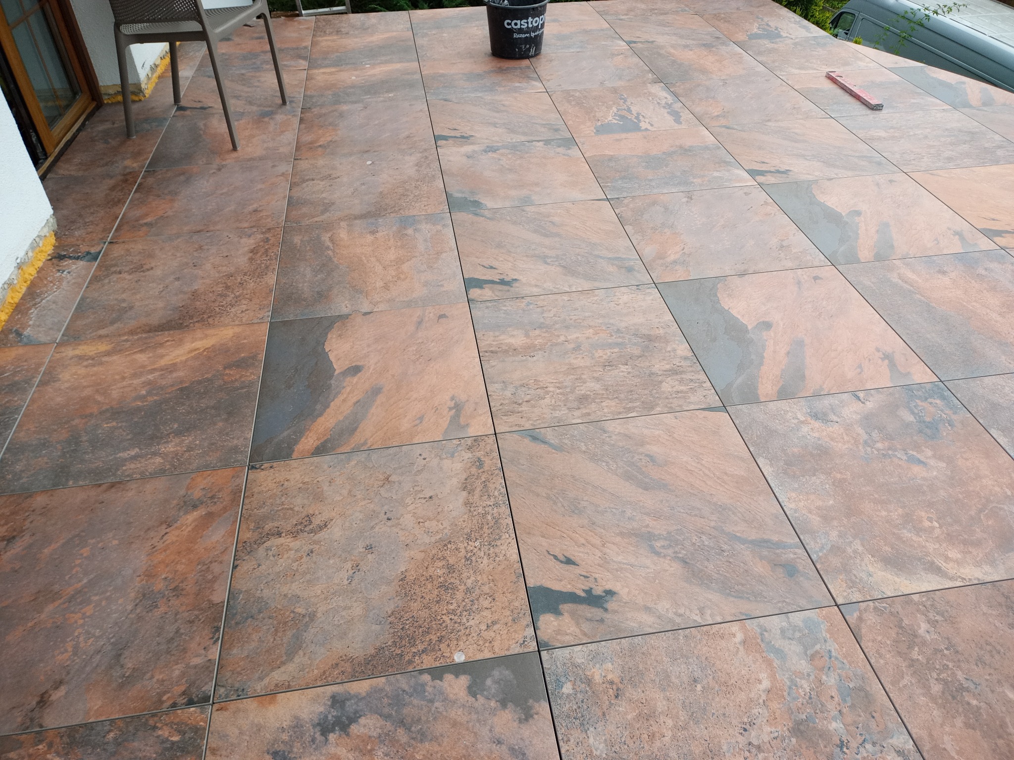 Wykończony taras z płytek imitujących kamień w odcieniach brązu i szarości. Widoczny kubeł z zaprawą i poziomica. Częściowo widoczne krzesło i fragment elewacji.