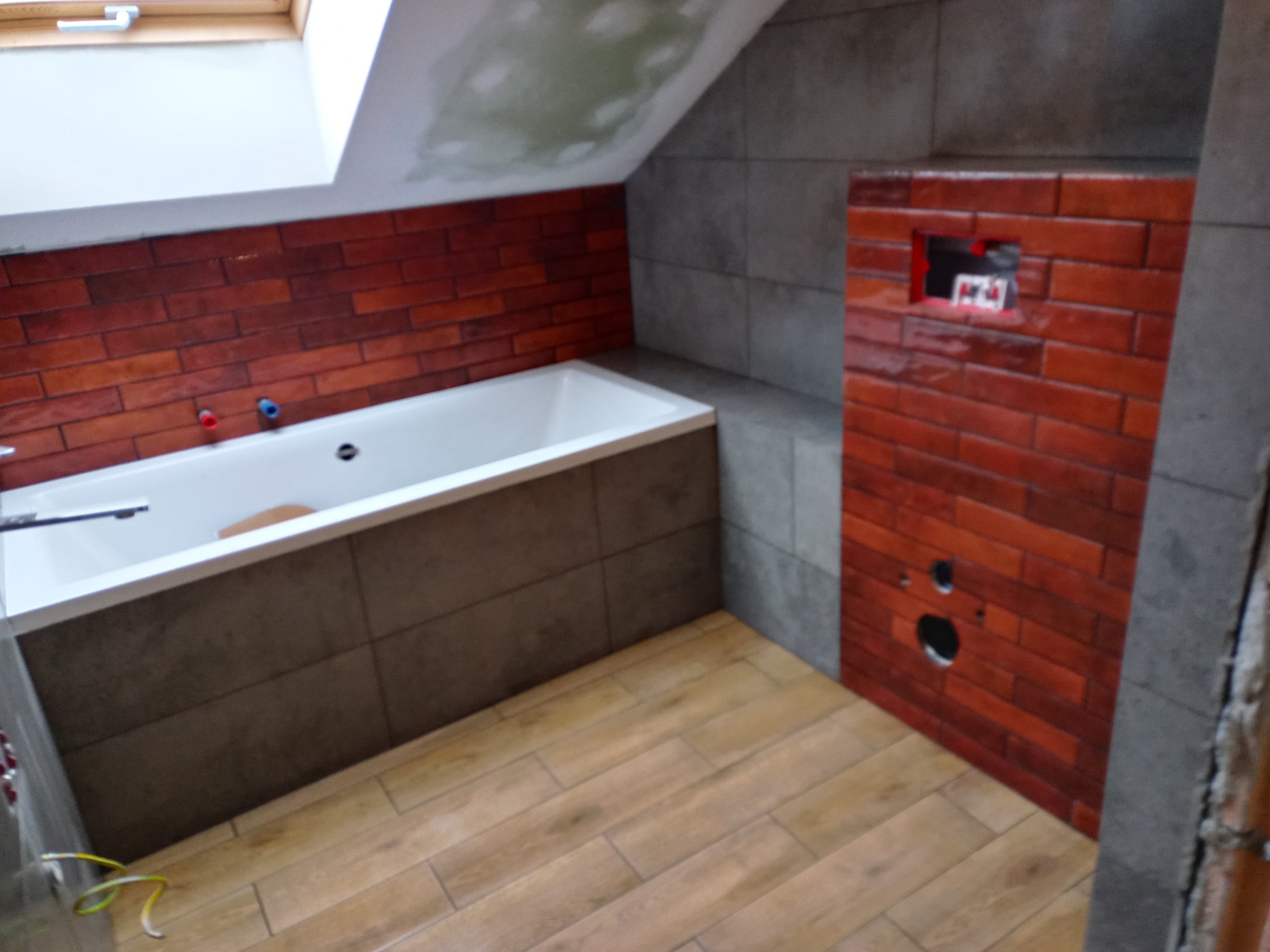 Ukończona łazienka na poddaszu: wanna obudowana szarą płytką, ściana z czerwonej cegły, drewnopodobna podłoga. Widoczne otwory na armaturę i okno dachowe.