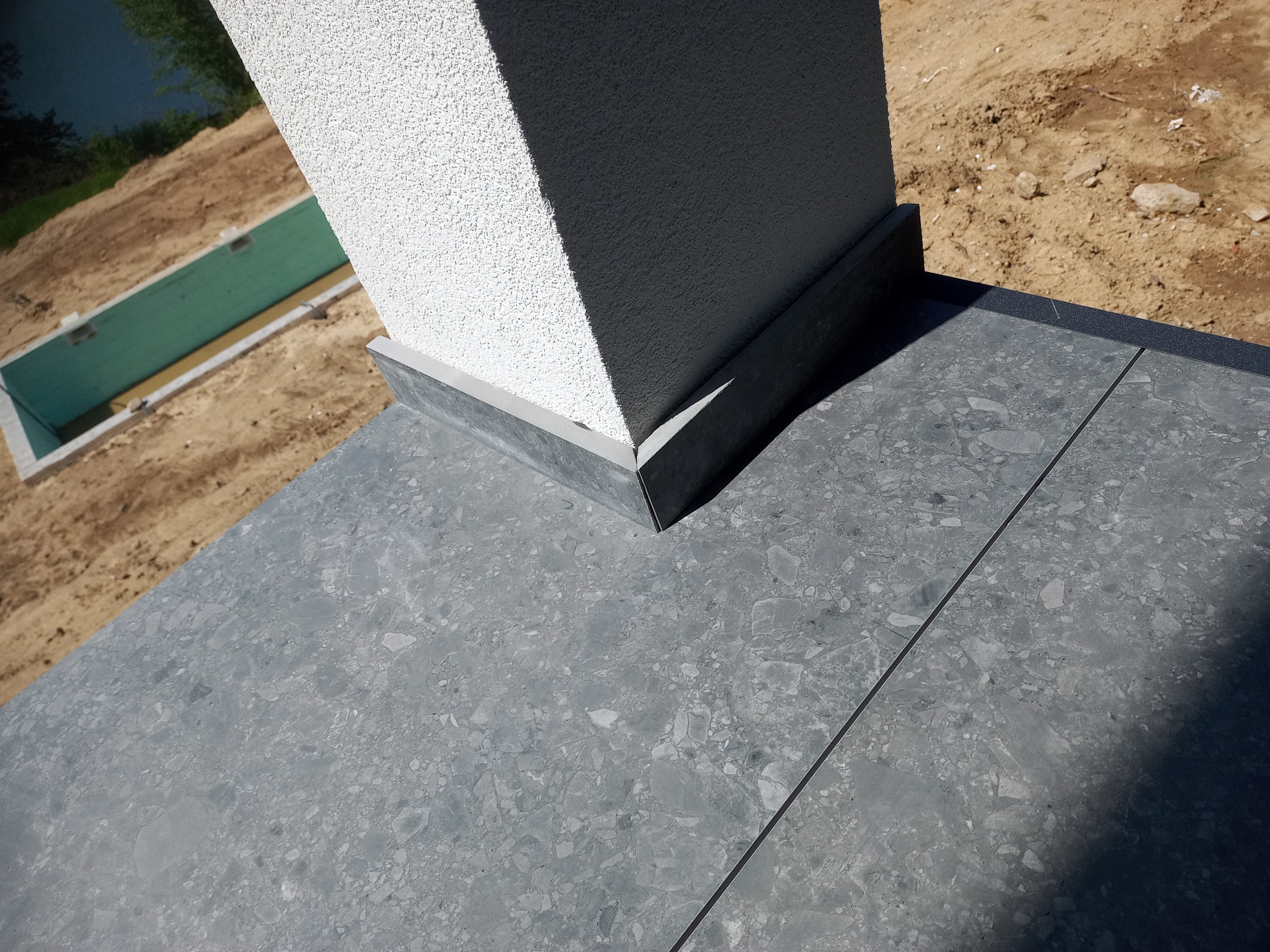 Szara, kamienna posadzka tarasu z fugami, przylegająca do otynkowanej kolumny. W tle widoczny fragment terenu z zielonymi elementami konstrukcyjnymi.