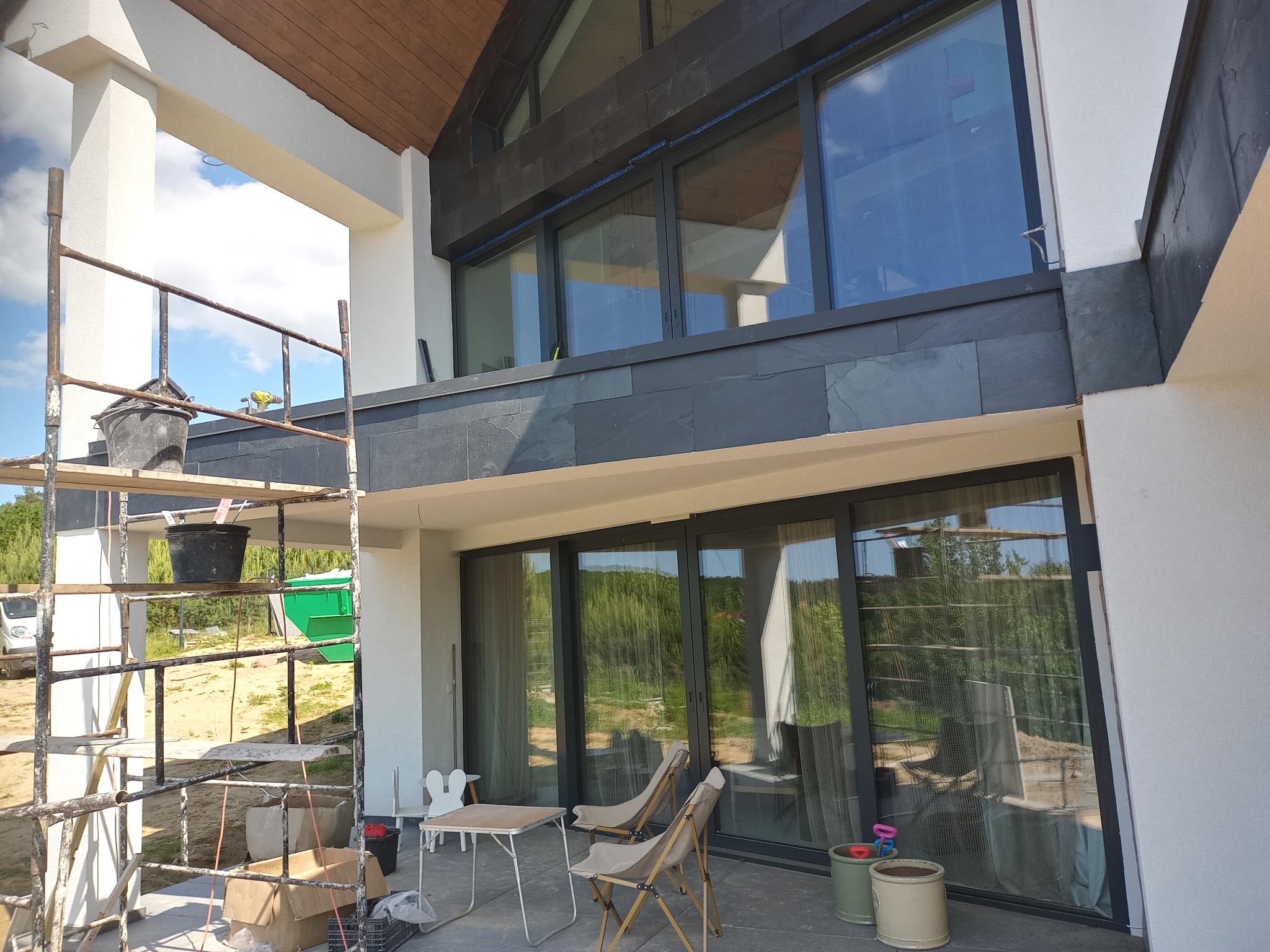 Nowoczesny dom z szarą elewacją i dużymi oknami, widok z rusztowania. Elementy wykończeniowe z kamienia. Na tarasie stolik i krzesła.