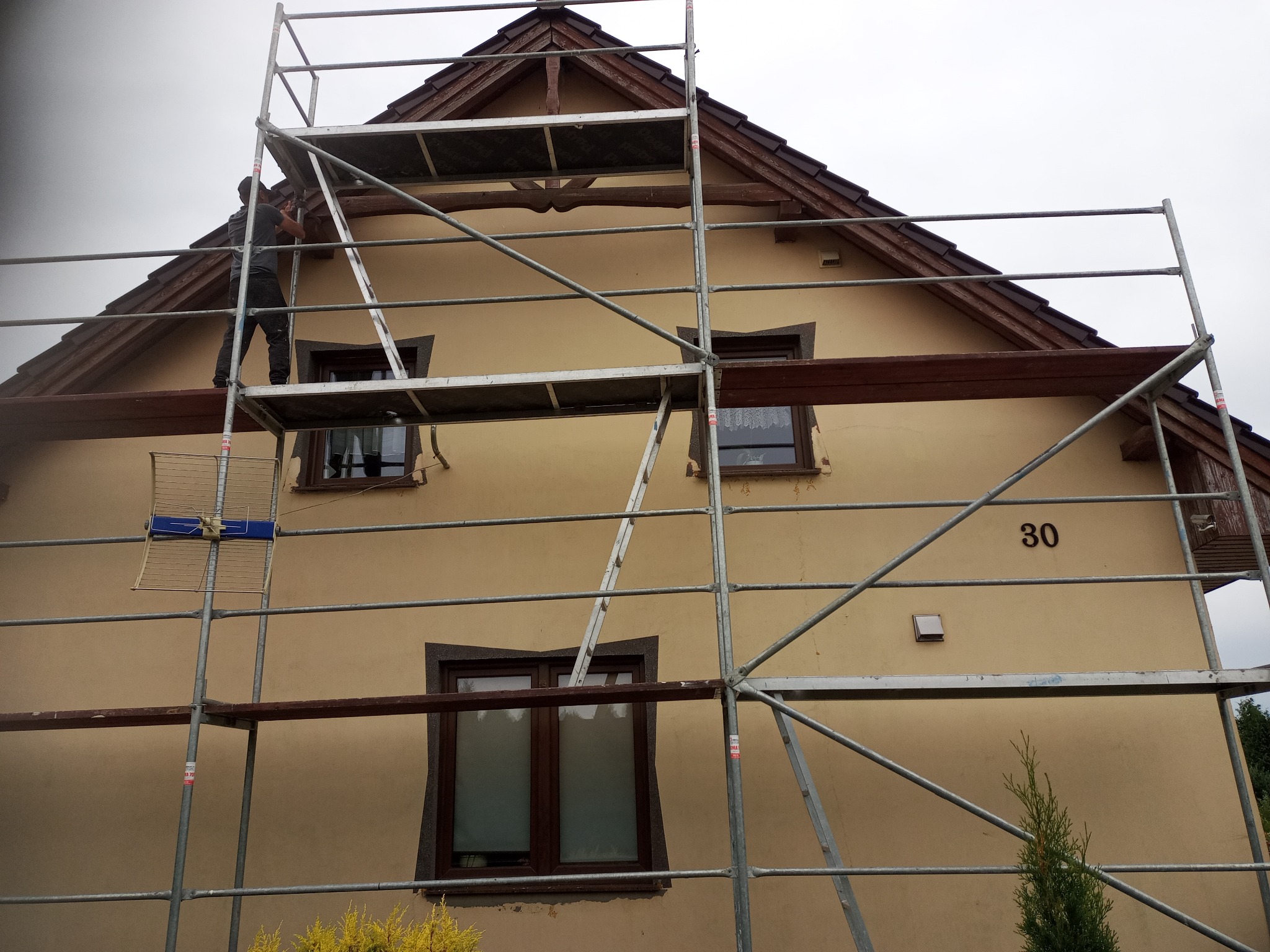 Praca na rusztowaniu przy elewacji domu z numerem 30. Widoczny pracownik przy dachu, okna z ciemnymi ramami i beżowa elewacja. Ujęcie z dołu.