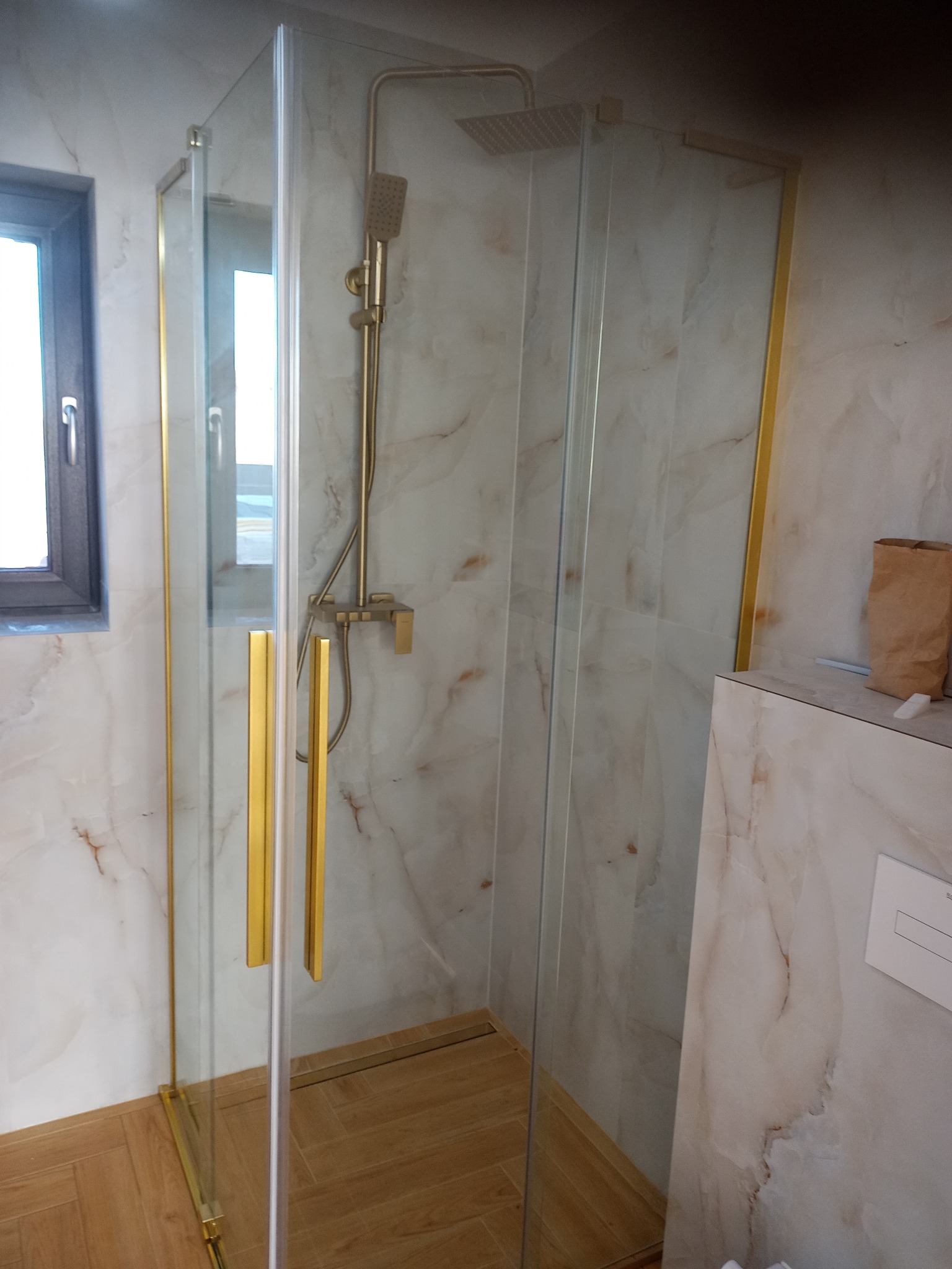 Nowoczesna kabina prysznicowa z złotymi detalami, marmurowe płytki na ścianach, drewnopodobna podłoga. Widok na okno z boku kabiny.