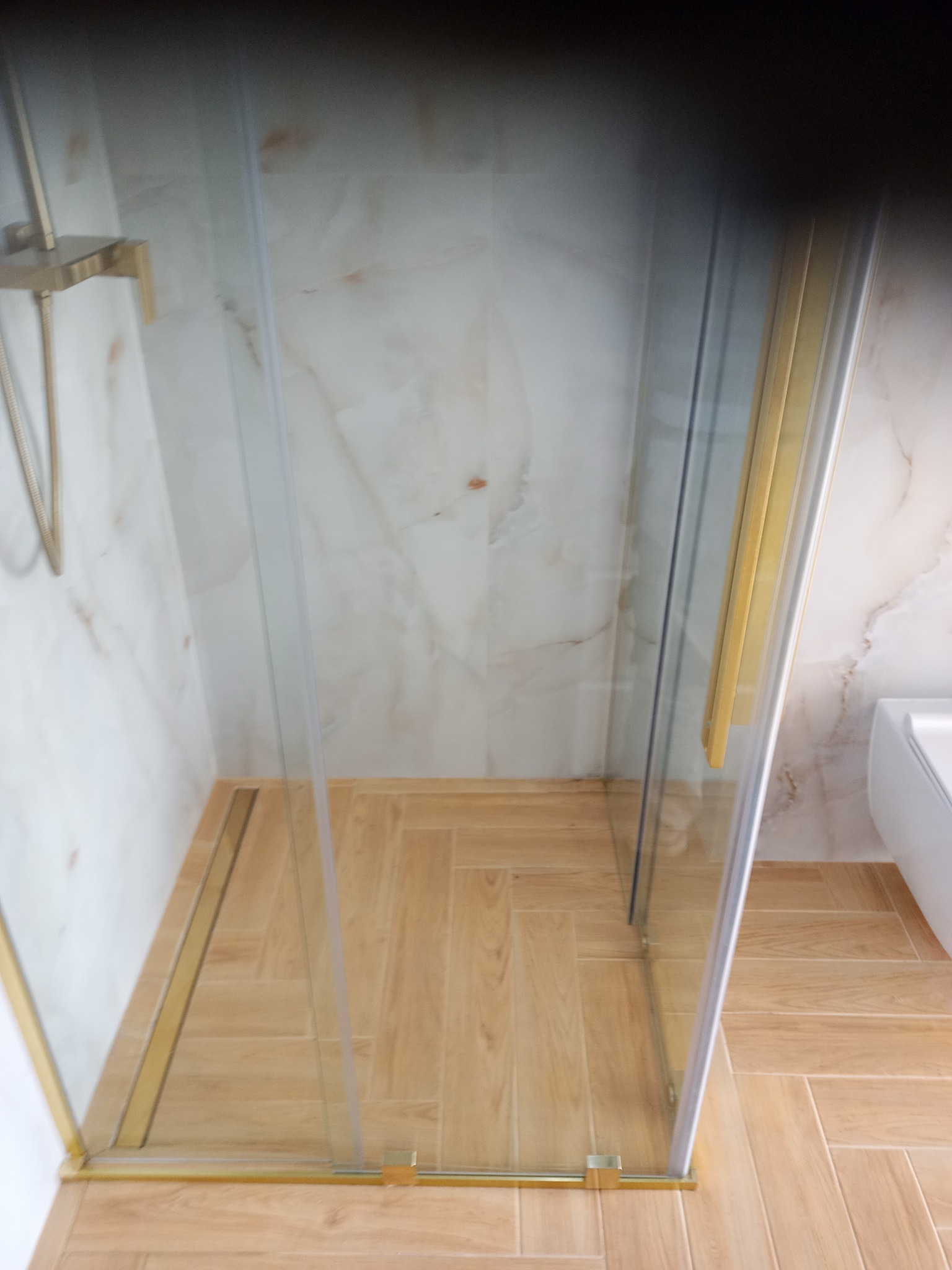 Nowoczesna kabina prysznicowa z złotymi elementami wykończeniowymi, jasne płytki imitujące marmur na ścianach i drewnopodobna podłoga w jodełkę.