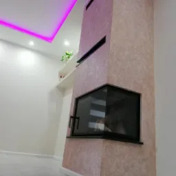 Otynkowany kominek narożny z czarną obudową, widoczny z boku, na tle jasnych ścian i podłogi z płytek, z podświetleniem LED na suficie.