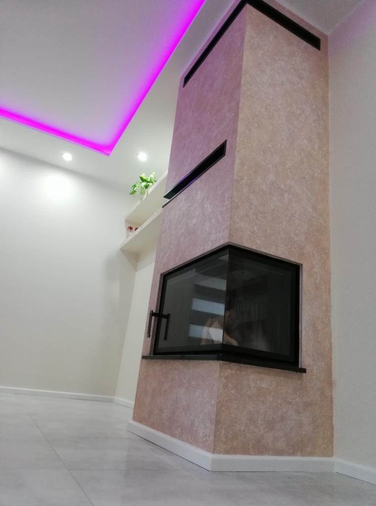 Otynkowany kominek narożny z czarną obudową, widoczny z boku, na tle jasnych ścian i podłogi z płytek, z podświetleniem LED na suficie.