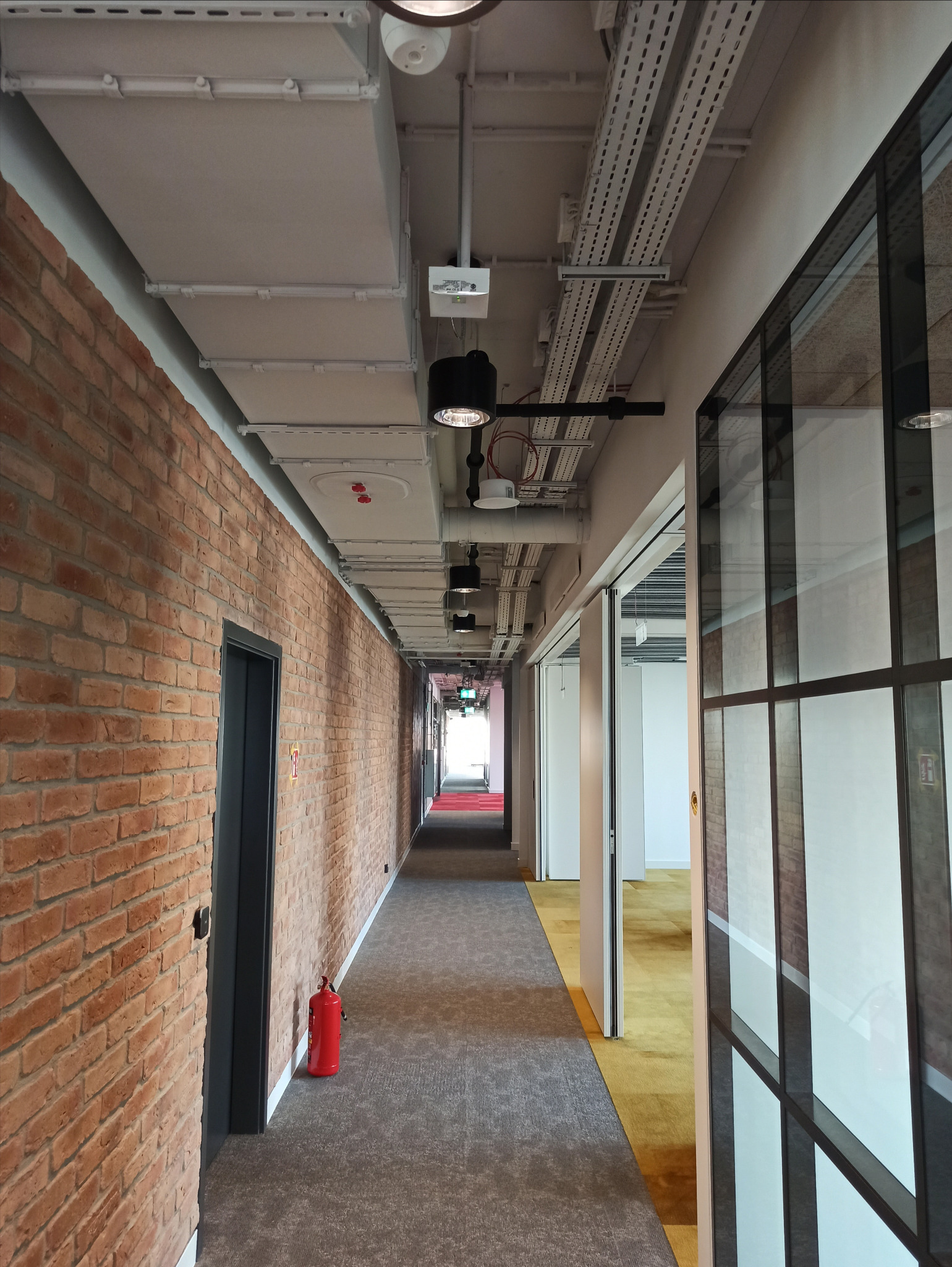 Długi korytarz biurowy z ceglaną ścianą, szarym dywanem i widocznymi instalacjami pod sufitem, oświetlony czarnymi lampami punktowymi.