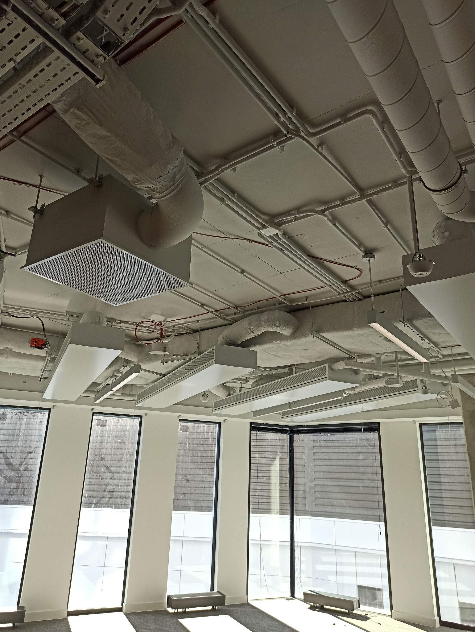 Wnętrze biura z widocznymi instalacjami pod sufitem: rurami wentylacyjnymi, przewodami elektrycznymi i lampami sufitowymi, świeżo po malowaniu na biało.