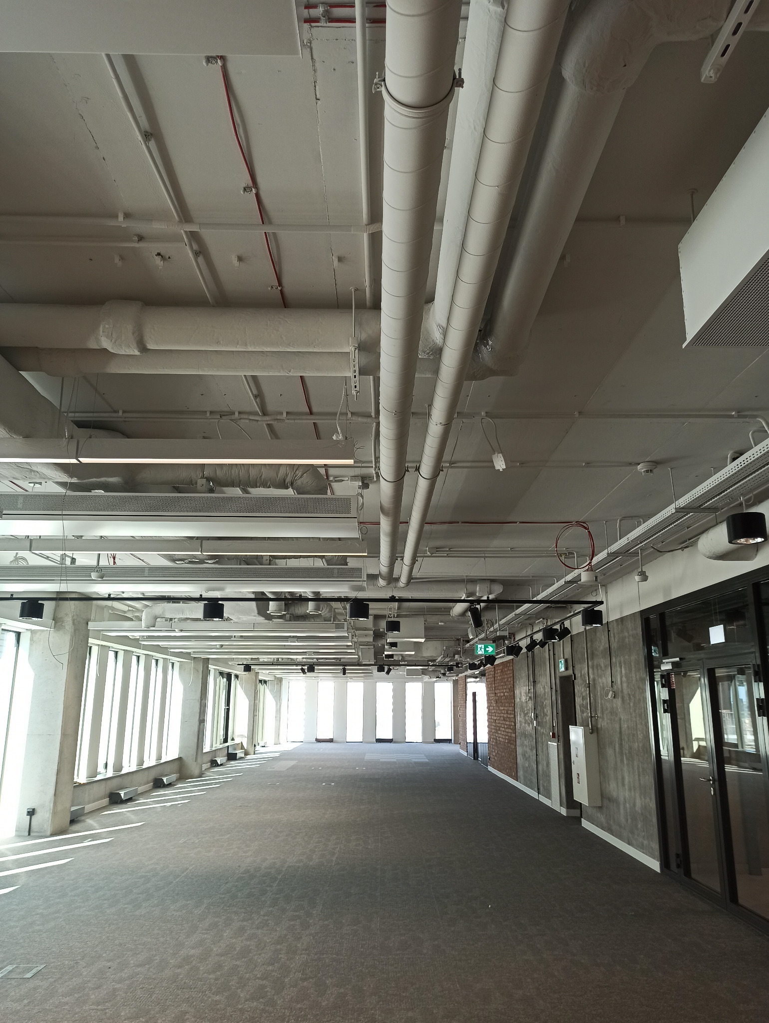 Przestronne wnętrze biurowe z widocznymi instalacjami pod sufitem, szarą wykładziną dywanową i dużymi oknami wpuszczającymi naturalne światło, ukazujące przygotowanie do aranżacji.