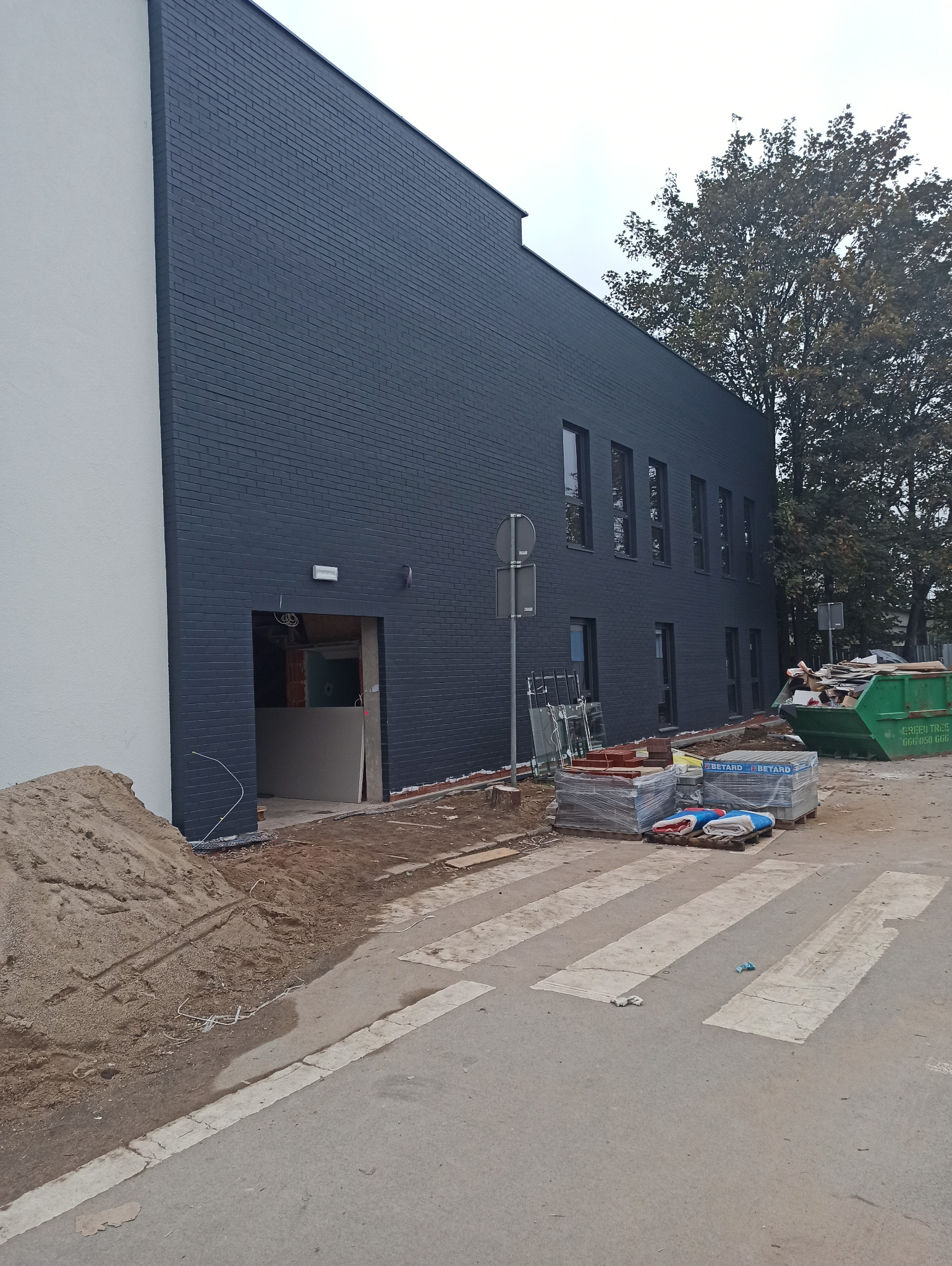 Budynek biurowy z pomalowaną na ciemny kolor ceglaną elewacją, widoczne okna, wejście w stanie surowym, obok sterta piasku i materiały budowlane na paletach.