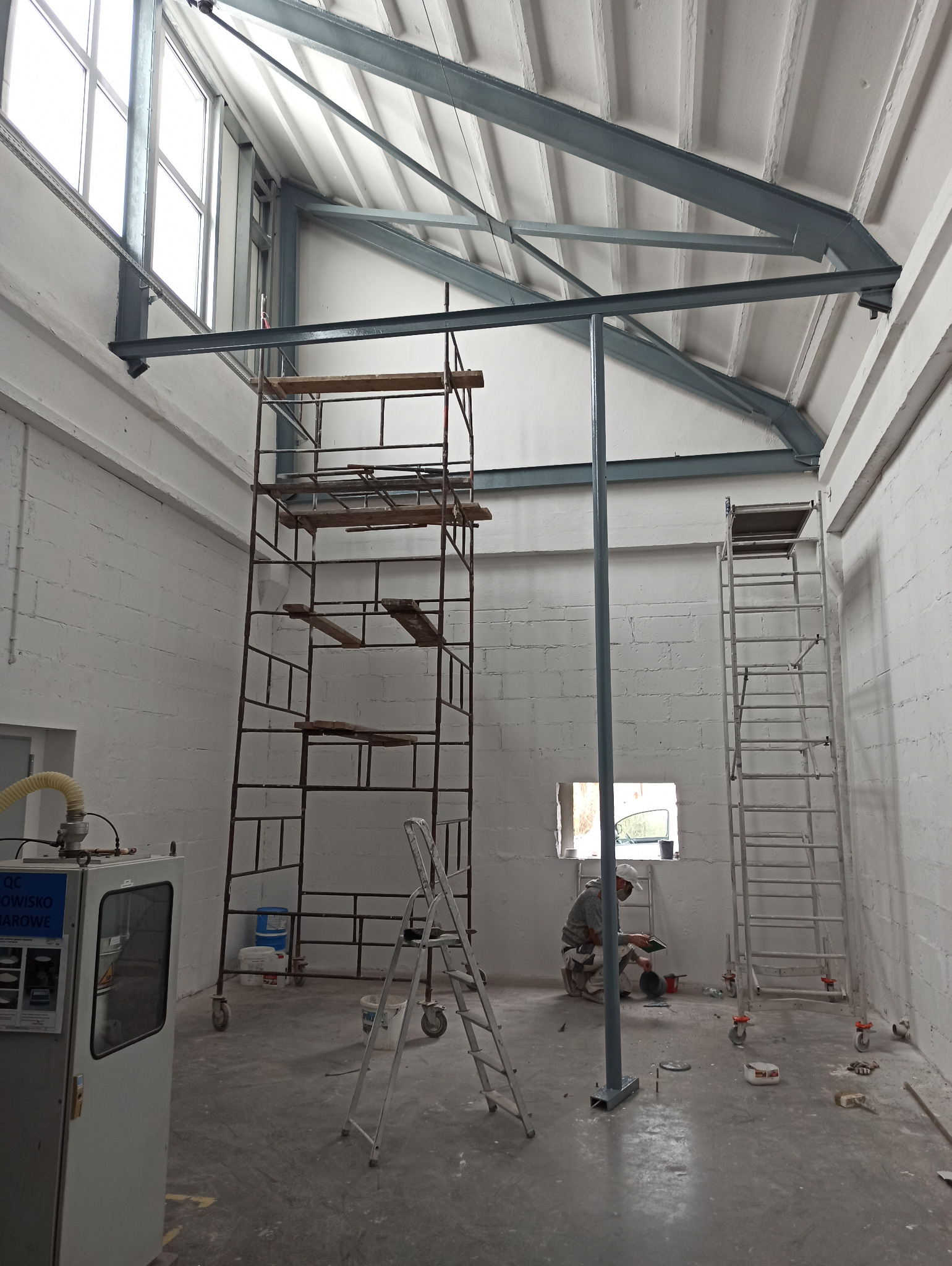 Wnętrze hali przemysłowej w trakcie prac wykończeniowych, widoczne rusztowania, drabina i pracownik przygotowujący farbę pod oknem, ściany pomalowane na biało