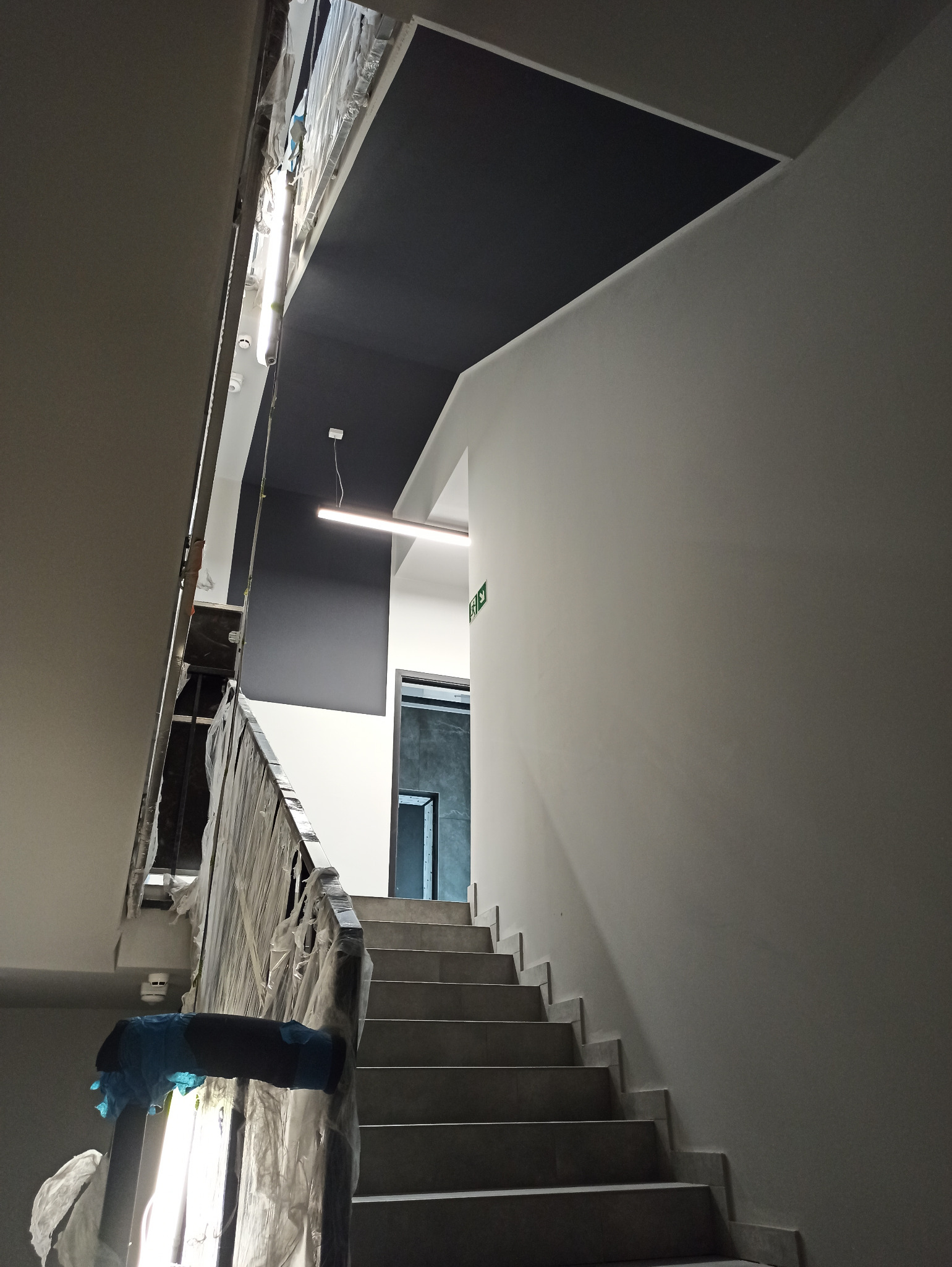 Klatka schodowa w biurze w trakcie prac wykończeniowych, ściany pomalowane na biało, sufit częściowo na czarno, zabezpieczona folią barierka schodów.