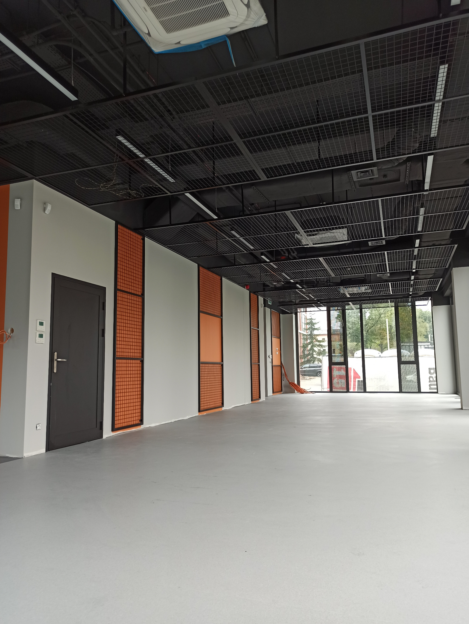 Wnętrze biura po remoncie, widoczne szare ściany z pomarańczowymi panelami, szara podłoga oraz czarny sufit z kratownicą i instalacjami.