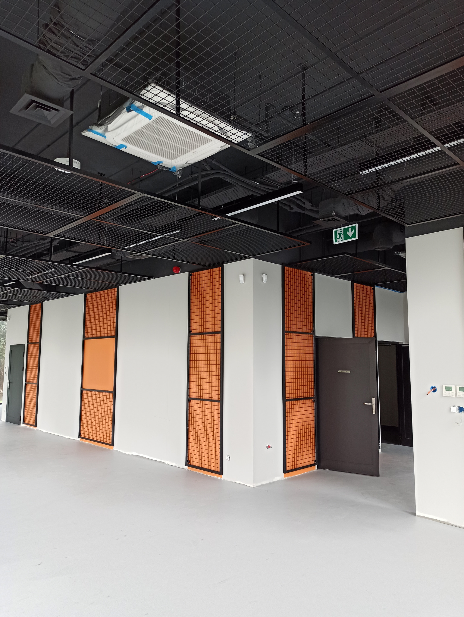 Wnętrze biurowe z białymi ścianami, pomarańczowymi panelami z siatki i czarną kratownicą sufitową, klimatyzator zabezpieczony folią, szare drzwi.