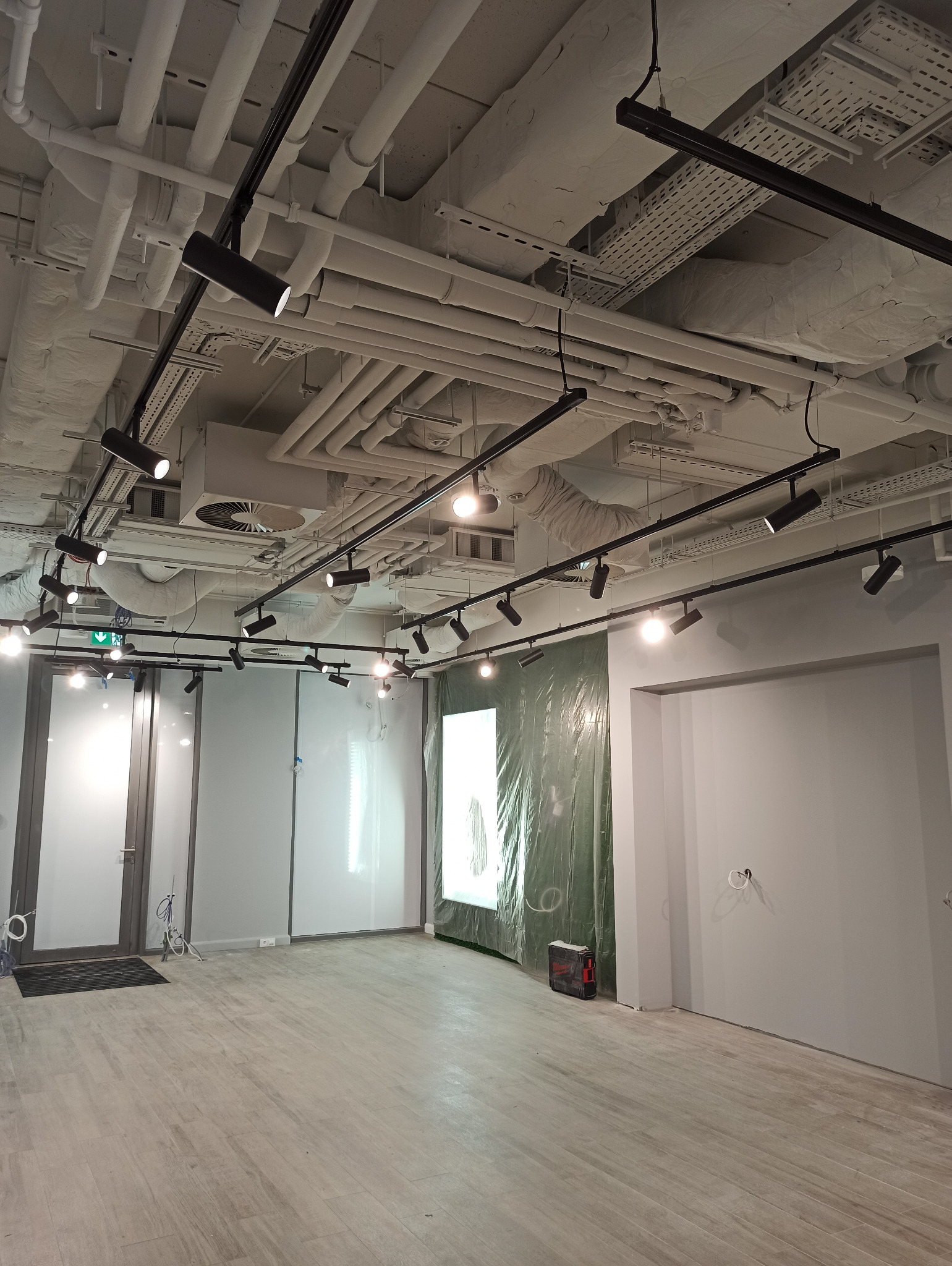 Wnętrze biura w trakcie prac wykończeniowych, widoczne zabezpieczone folią okno, surowy sufit z instalacjami i oświetleniem szynowym, oraz przygotowane do malowania ściany.