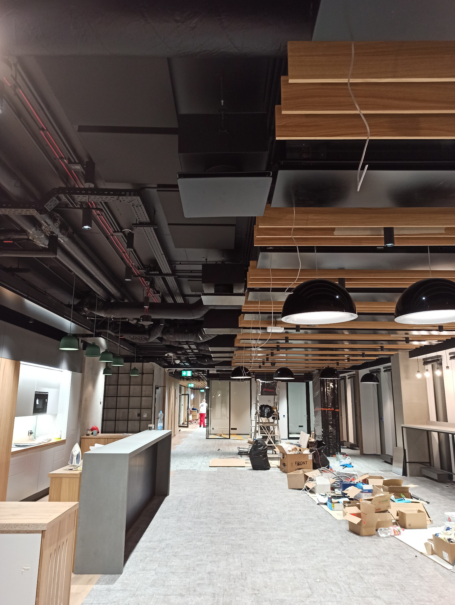 Wnętrze biura w trakcie prac wykończeniowych: widoczny surowy betonowy bar, podwieszany sufit z instalacjami, rozłożone kartony i narzędzia na podłodze, oraz fragment wyposażonej kuchni w tle.