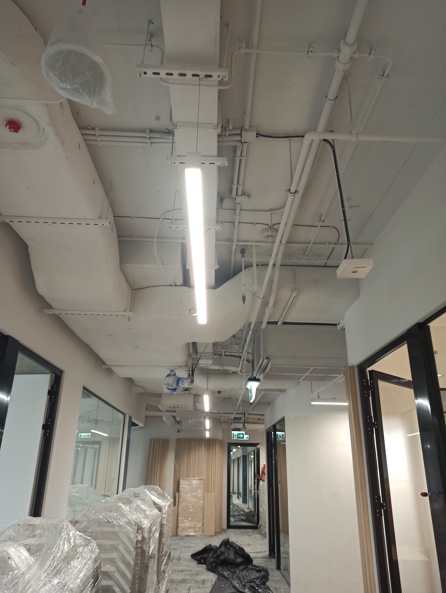 Wnętrze biura w trakcie prac wykończeniowych, widoczne instalacje pod sufitem, zabezpieczone meble folią i elementy ścian do montażu.