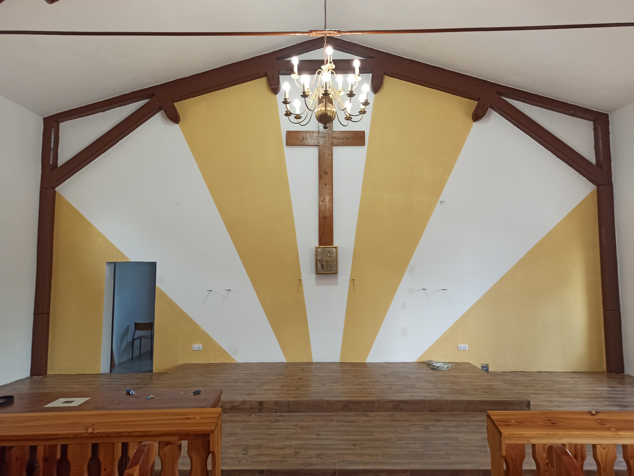 Wnętrze po odświeżeniu: ściana z geometrycznym wzorem w odcieniach bieli i żółci, drewniany krzyż na ścianie, ozdobny żyrandol, drewniane elementy konstrukcyjne pod sufitem, widoczne ślady po...