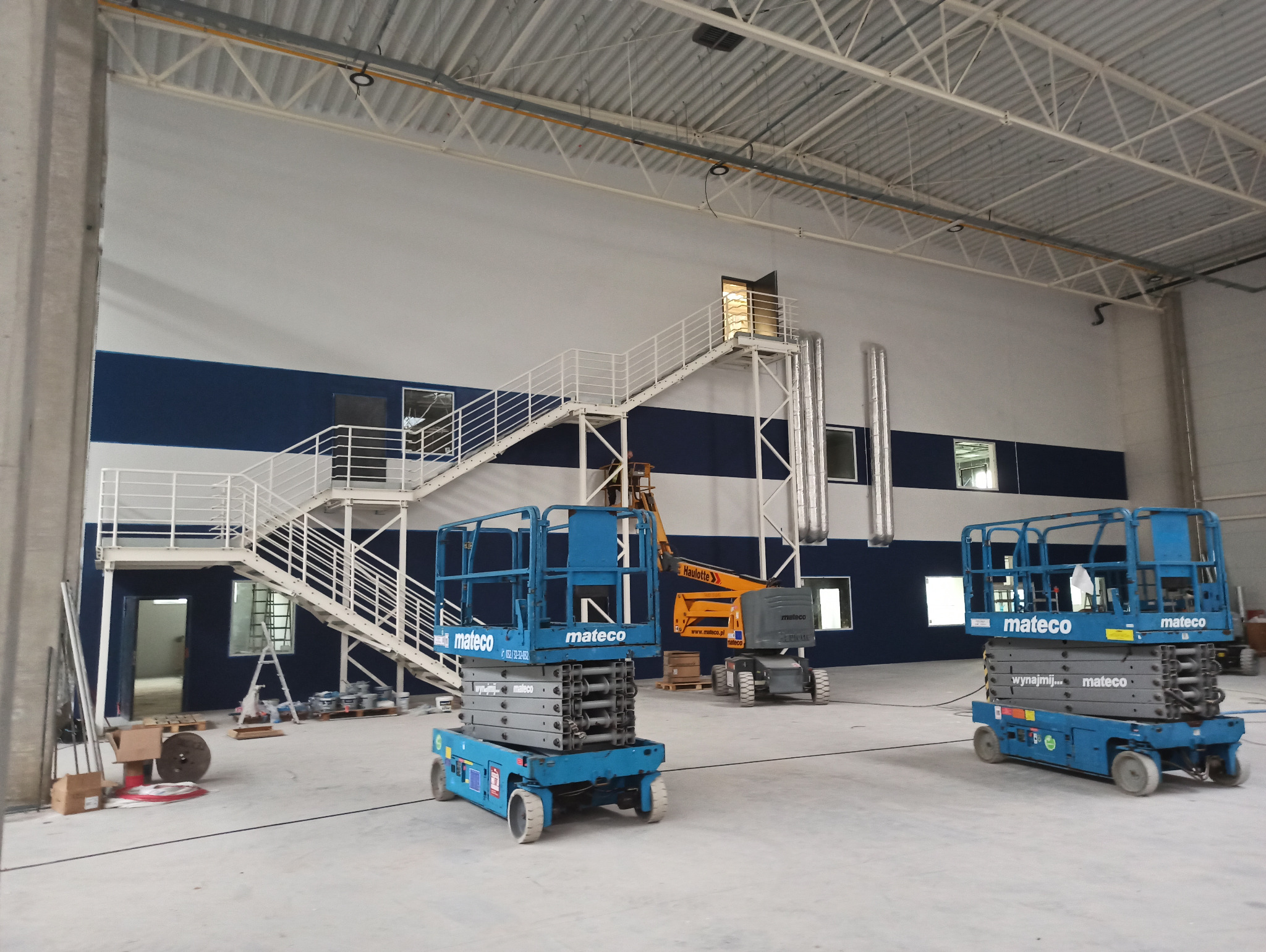 Wnętrze hali przemysłowej w trakcie malowania: widoczne podnośniki koszowe Mateco, pomalowane pasy biało-granatowe na ścianach, metalowe schody z białymi barierkami i rozstawiona drabina malarska...